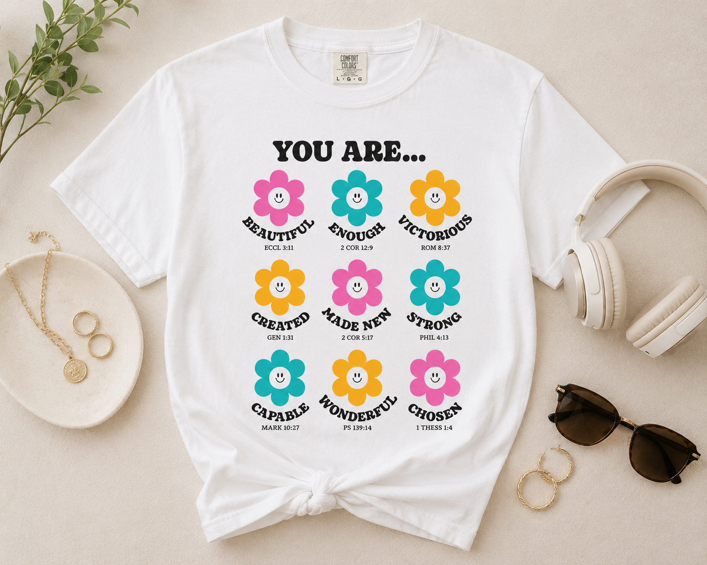 White Christian affirmation flower t-shirt flat lay with gold jewelry, cream headphones, sunglasses, and neutral modern lifestyle styling on a beige background