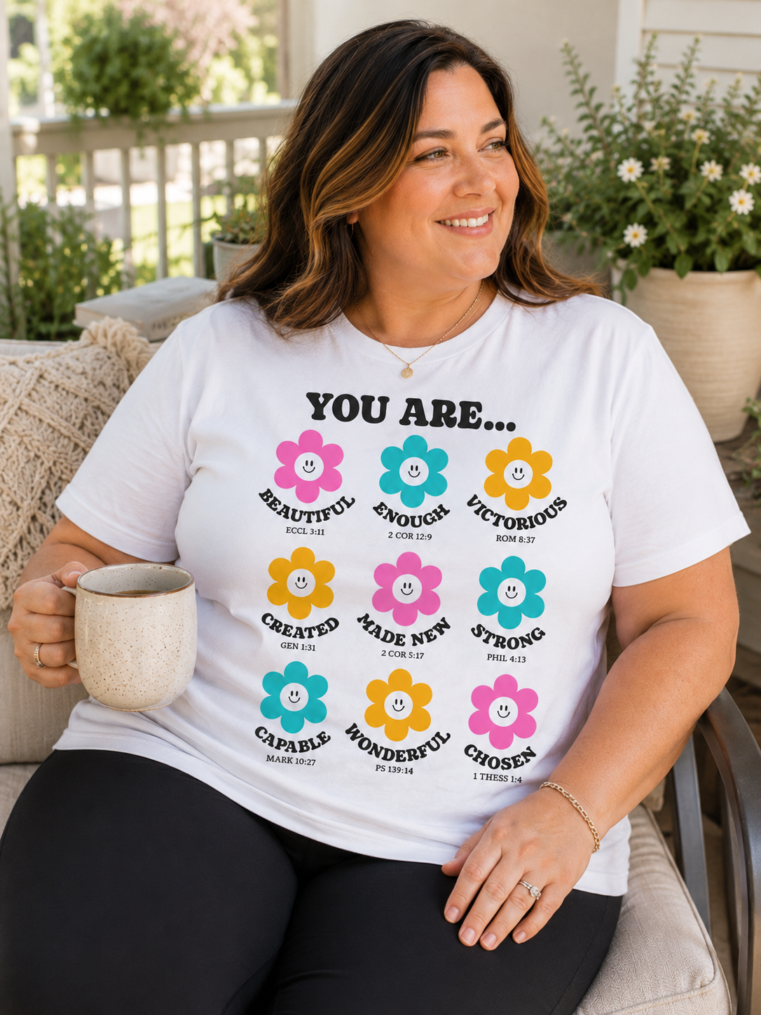 Plus size woman wearing white Christian affirmation flower t-shirt with black leggings while relaxing outdoors on a cozy porch with coffee mug and soft natural light