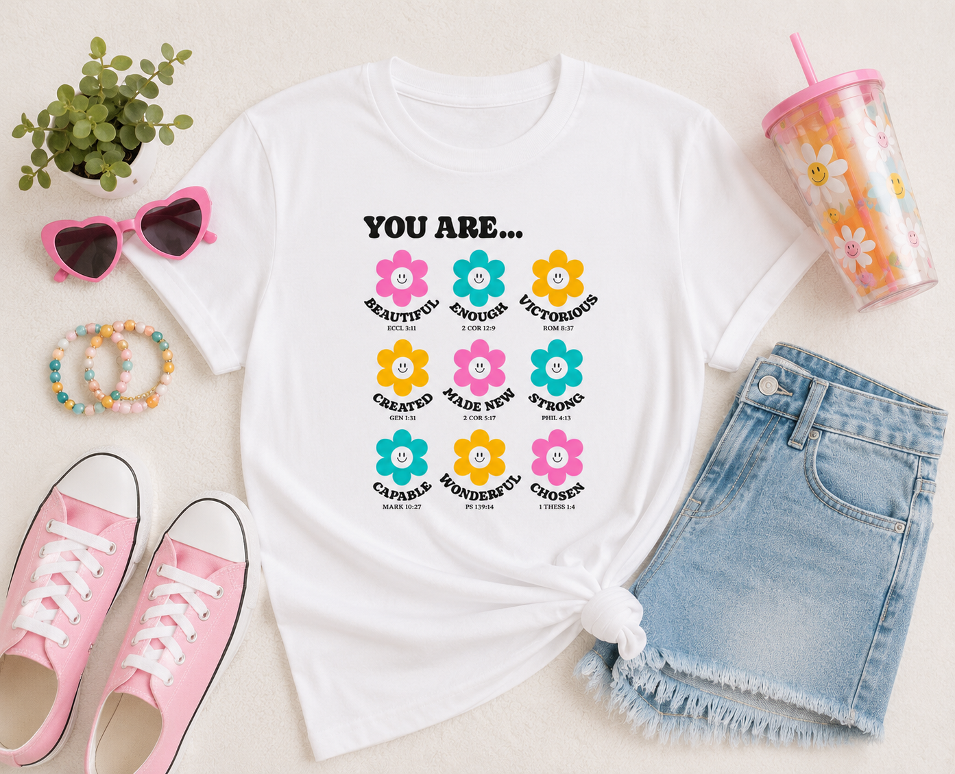 Flat lay mockup of a white “You Are…” Christian youth graphic t-shirt styled with pastel accessories, pink sneakers, denim shorts, and playful modern decor in a bright youthful aesthetic.