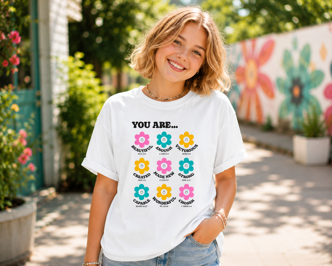 Tween girl with short blonde hair wearing a white “You Are…” Christian youth graphic t-shirt outdoors in a bright colorful setting with a modern fun youthful vibe.